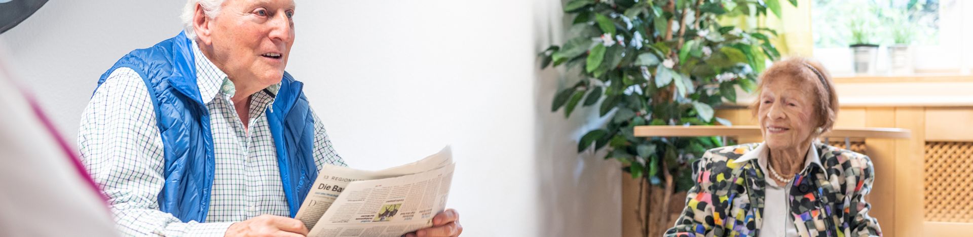 Älterer Mann liest Zeitung und ältere Frau im Rollstuhl hört zu